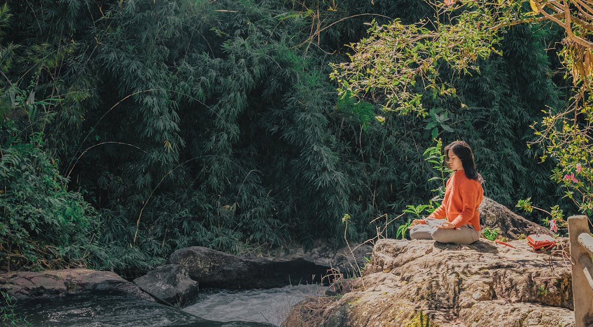 Woman meditating by a serene riverside surrounded by lush greenery on a sunny day.